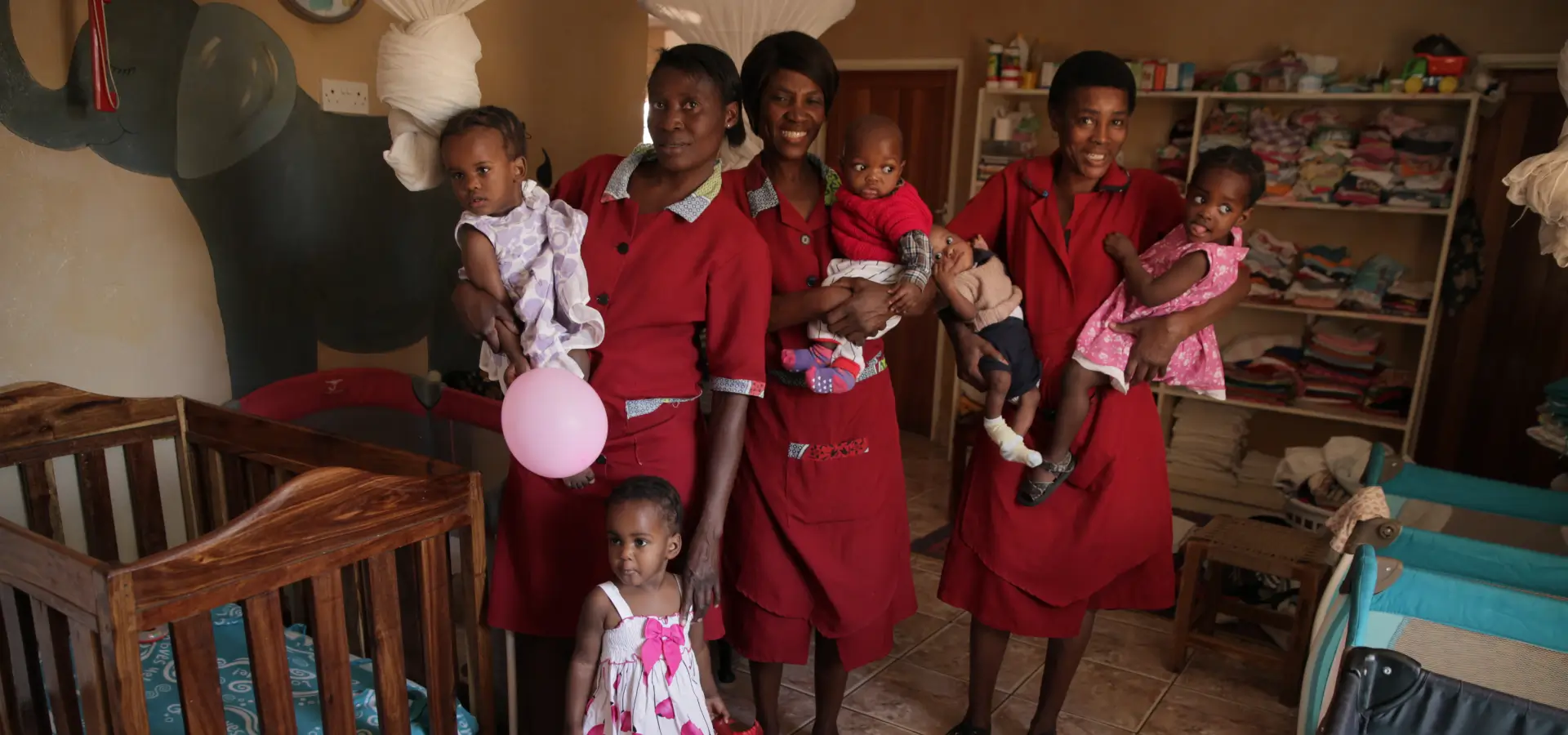 Three women in red dresses holding babies and balloons indoors.